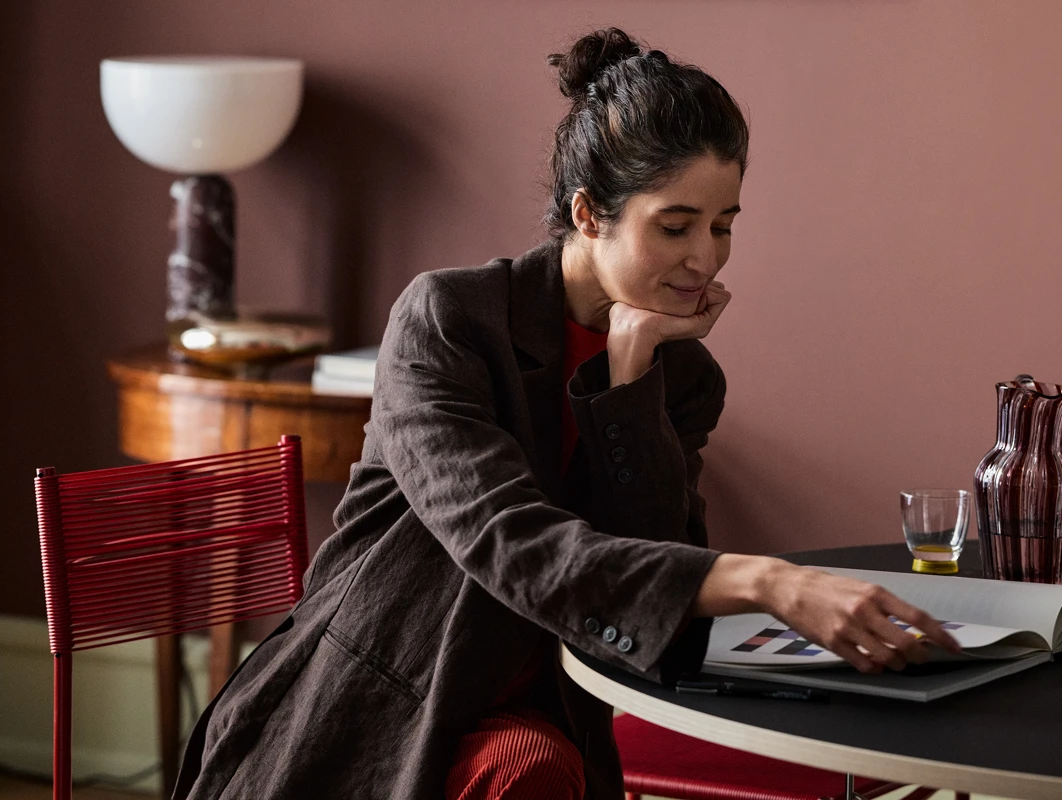 Woman in a brown jacket reading a book while seated at a table in a warmly decorated room, featuring a red chair, lamp, and glassware